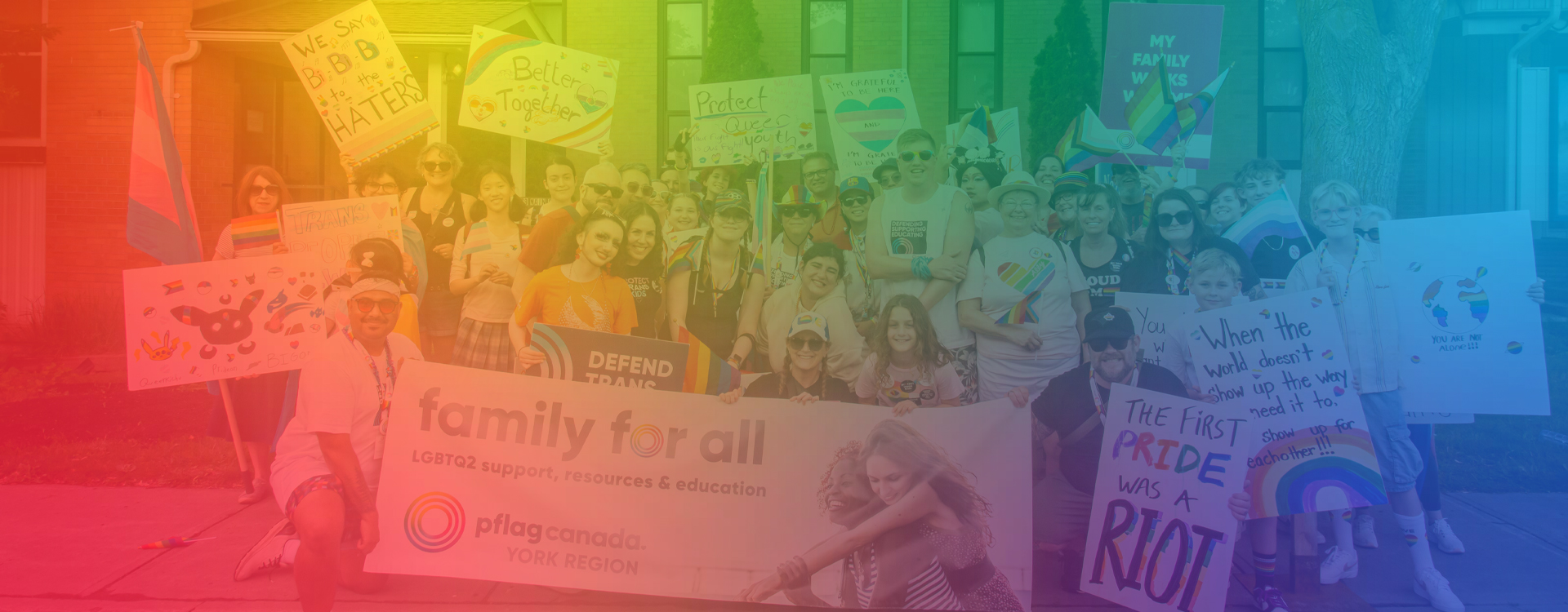 Group of Pride participants holding rainbow flags, LGBTQ2S+ signs, and a Pflag Canada York Region banner outside a building.